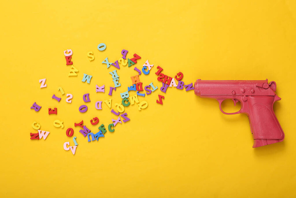 Pink pistol gun shooting letters on yellow background. Creative minimalism layout. Concept art. Flat lay. Top view - Photo, Image