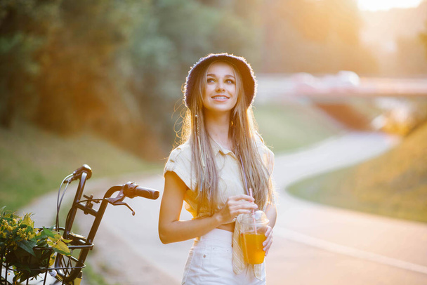 A happy girl in white shorts and a yellow blouse holds a glass of juice with a straw on the background of the road near the forest - Photo, Image