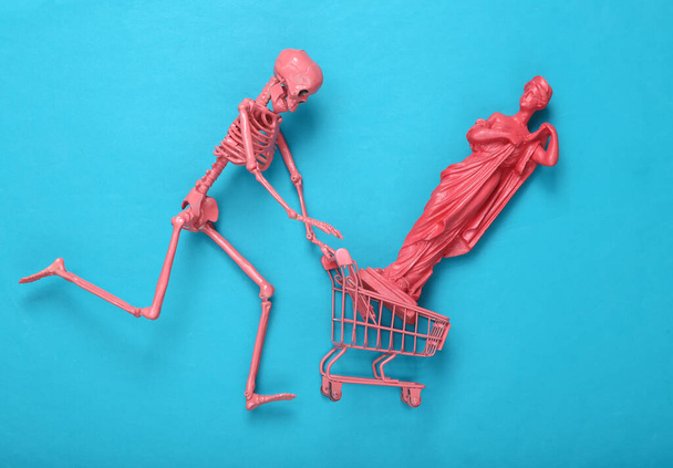 Mini růžový supermarket vozík, starožitná socha a kostra na modrém pozadí. Rozvržení Halloweenu minimalismu - Fotografie, Obrázek