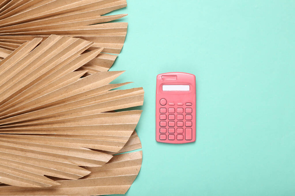 Creatieve indeling. Roze rekenmachine op blauwe achtergrond met droge palmbladeren. Plat gelegd. Bovenaanzicht - Foto, afbeelding