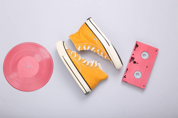 Creative retro 80s layout. Pink vinyl record, video cassette and sneakers on gray background. Minimalism. Flat lay. Top view. - Photo, Image