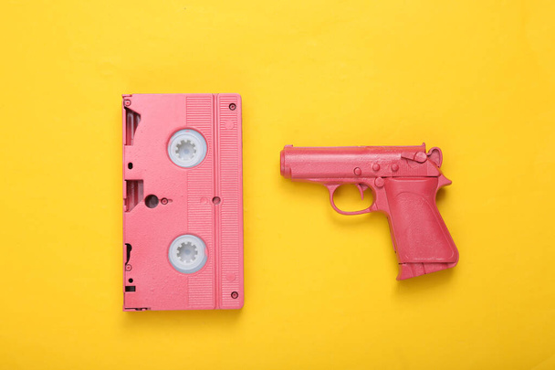 Roze videocassette en pistool op gele achtergrond. Creatieve indeling. Minimalisme. Plat gelegd. Bovenaanzicht. - Foto, afbeelding