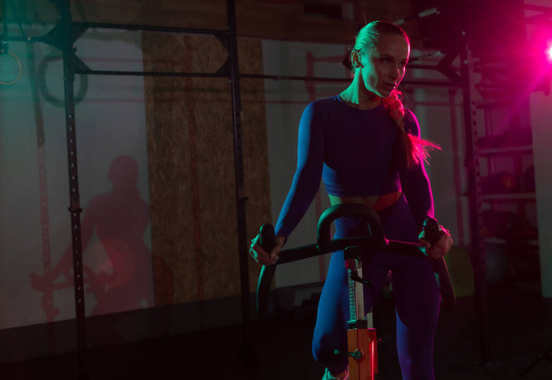 Krásná mladá fit žena dělá kardio na stacionárním kole v růžové zelené neon gradient světlo v tělocvičně. Sport, fitness, životní styl koncepce - Fotografie, Obrázek
