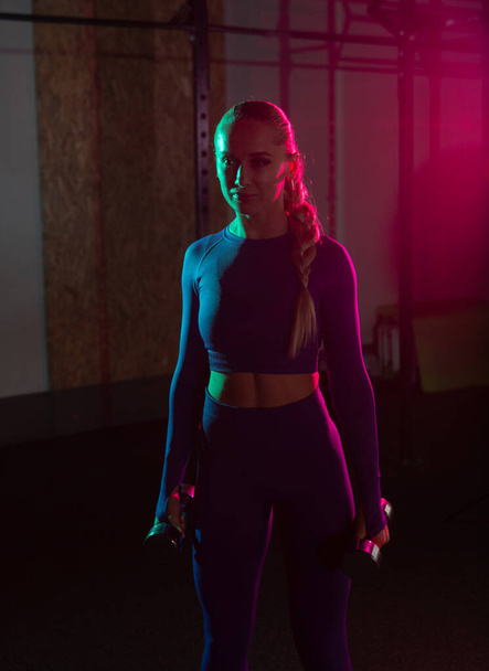 Young fit slim woman with dumbbells in her hands in pink green neon gradient light at gym. Sport, fitness, lifestyle concept - Φωτογραφία, εικόνα
