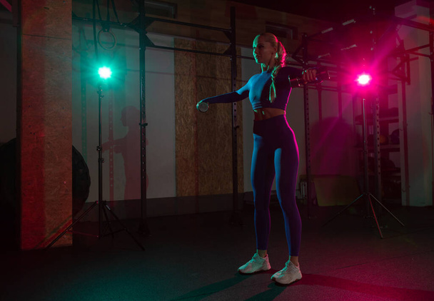 Молодая стройная женщина тренирует мышцы плеч с гантелями в руках в розово-зеленом неоновом градиентном свете в спортзале. Спорт, фитнес, стиль жизни - Фото, изображение