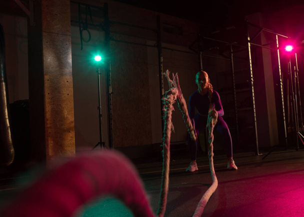 Атлетическая сильная женщина делает несколько упражнений с боевой веревкой в современном спортзале. Розовый зелёный неоновый свет. Функциональное обучение - Фото, изображение