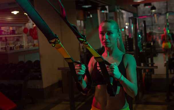 Spor giyim trenlerindeki zinde kadın spor salonunda kablo dirençli trx sistemiyle. Pembe yeşil neon ışık  - Fotoğraf, Görsel