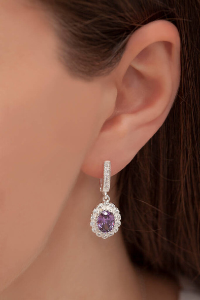 La mujer de piel blanca con pelo castaño rojizo posa con un pendiente de diamantes de color púrpura en forma de gota. - Foto, Imagen