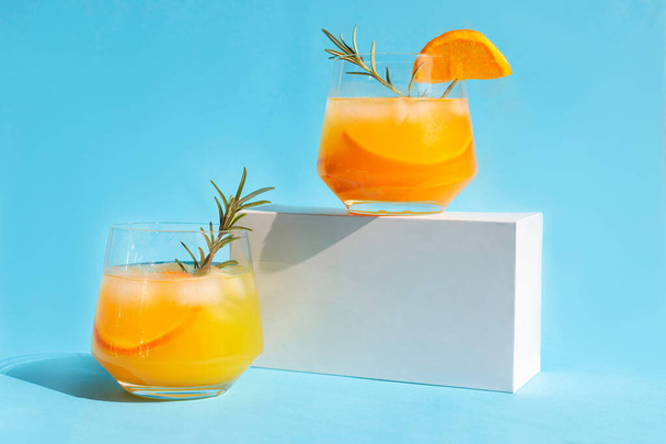 Lemonade, orange cocktail, refreshing summer drink with ice and orange slices in a glass with a sprig of rosemary on a white podium. Trending drinks. - Photo, Image