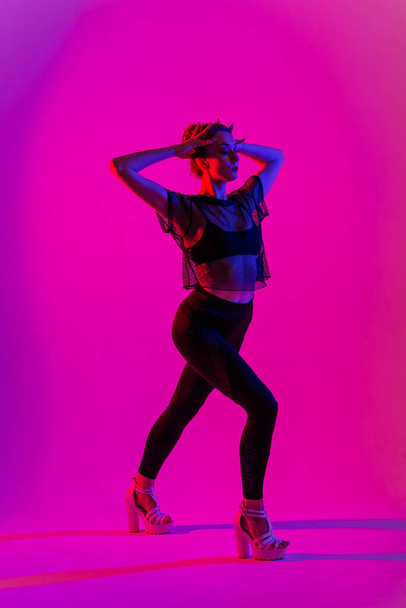 Beautiful female dancer wearing black and high heels while performing pole dance tricks, neon - Photo, image