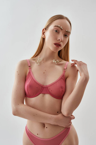Slim young caucasian female model with flowers in her eyebrows and on the clavicles wearing pink transparent underwear standing, posing isolated over light gray background - 写真・画像