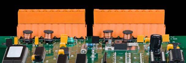 Detalle panorámico de componentes electrónicos en placa de circuito verde sobre fondo negro. Primer plano de virutas, bobinas cilíndricas con alambre de cobre y transistores o condensadores. Dos bloques terminales naranjas. - Foto, imagen
