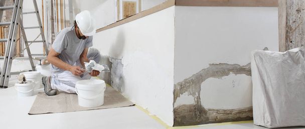 штукатурка людина на роботі, візьміть розчин з відра шпателем для штукатурки стіни інтер'єру будівельного будинку і носіння шолома, панорамне зображення
 - Фото, зображення