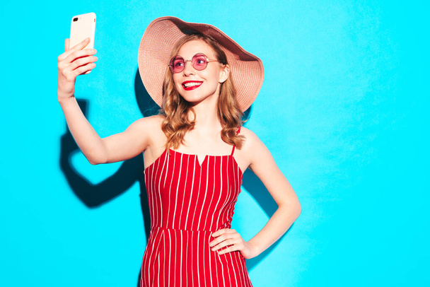 Young beautiful smiling female in trendy summer red hipster overalls. Sexy carefree woman posing near blue wall in studio. Positive model having fun in sunglasses and hat. Taking selfie photos - Φωτογραφία, εικόνα