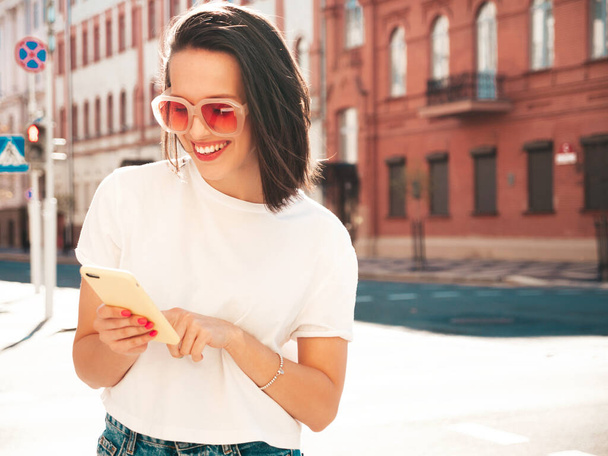 Young beautiful smiling hipster woman in trendy summer clothes. Sexy carefree woman posing on the street background in sunglasses. Positive model outdoors using smartphone apps, looking at screen - Фото, изображение