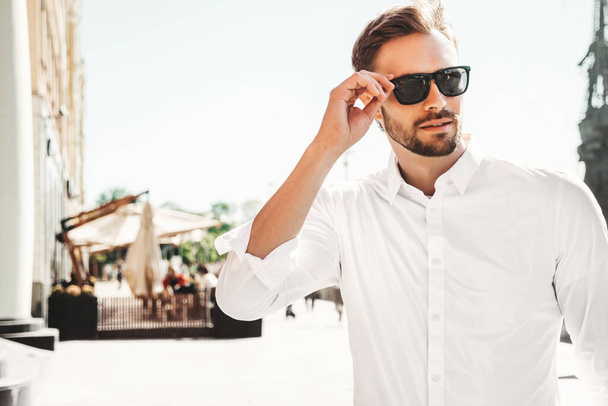 Portrait of handsome smiling stylish hipster lambersexual model.Modern man dressed in white shirt. Fashion male posing on the street background in sunglasses. Outdoors at sunset  - Photo, image