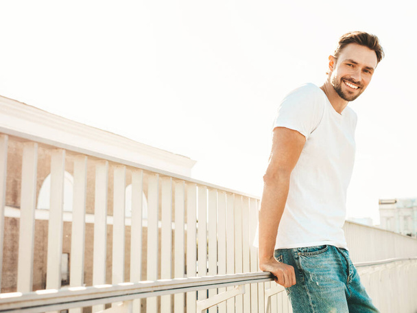 Portrait of handsome smiling  hipster lambersexual model.Stylish man dressed in white T-shirt and jeans. Fashion male posing on the street background near fence at the beach in sunglasses - Foto, Bild