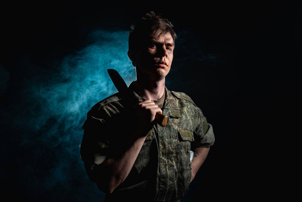 Portrait of a soldier with scars and combat coloring, holding a machete in the urns on a black background witch blue smoke - Photo, image
