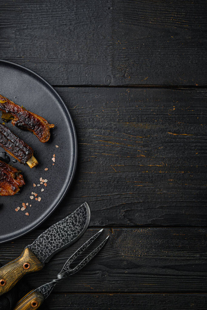 Grilled barbecue varkensvlees ribbetjes set, op bord, met barbecue mes en vlees vork, op zwarte houten tafel achtergrond, bovenaanzicht plat lay, met kopieerruimte voor tekst - Foto, afbeelding