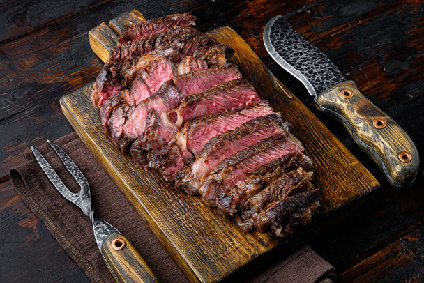 Соковита середня яловичина Ребро Стейк скибочки на дерев'яній дошці, з м'ясним ножем і виделкою, на старому темному дерев'яному фоні столу
 - Фото, зображення