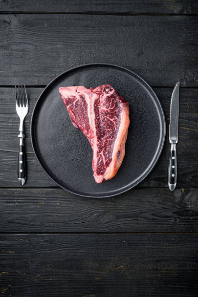 Roh frisches, trocken gereiftes Rindfleisch-T-Bone-Steak-Set, auf Teller, auf schwarzem Holztischhintergrund, von oben flach gelegt, mit Kopierplatz für Text - Foto, Bild