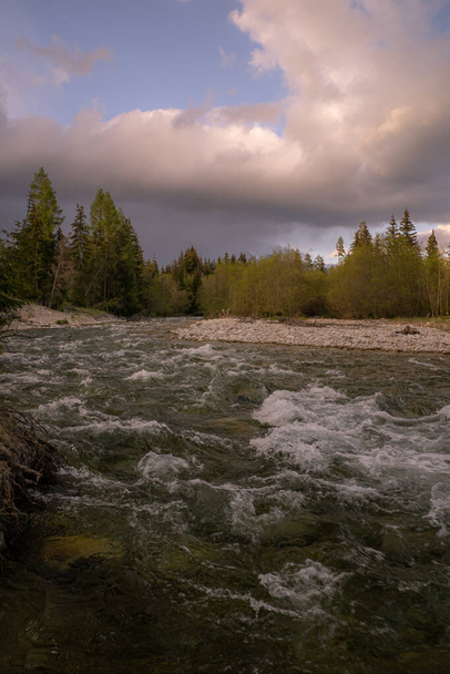 Ein stürmischer Gebirgsfluss fließt in ein Tal mit Felsen am Ufer und einem wolkenverhangenen Himmel darüber. - Foto, Bild