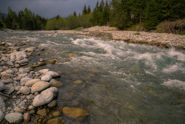 Une rivière de montagne orageuse coule dans une vallée avec des rochers sur les rives et un ciel nuageux au-dessus. - Photo, image