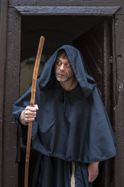 Portret van een bejaarde gebochelde monnik van 45-50 jaar met een baard in een zwart gewaad met een staf die uit de deur van een klooster of kerk komt, wegkijkend. - Foto, afbeelding