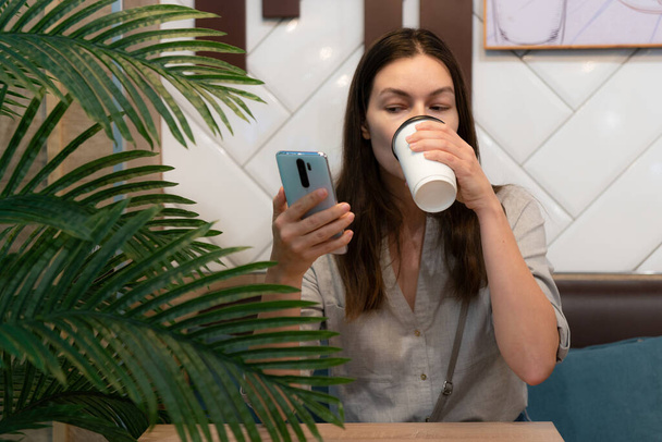 Молода жінка сидить за столом у кафе і спілкується в Інтернеті за допомогою мобільних додатків., - Фото, зображення