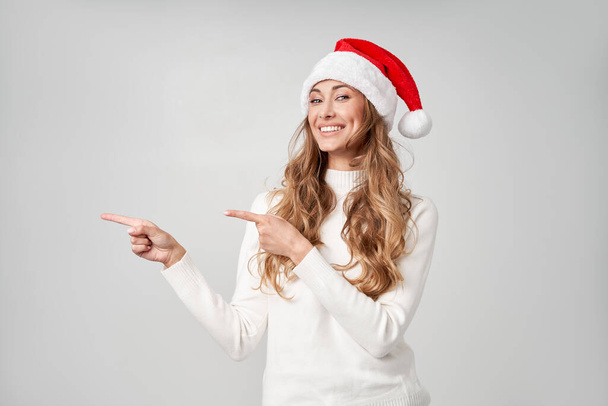 Woman christmas Santa Hat sweater white studio background Beautiful caucasian female curly hair portrait Happy person positive emotion Holiday concept Shows finger on side cope space for advertise - Фото, зображення