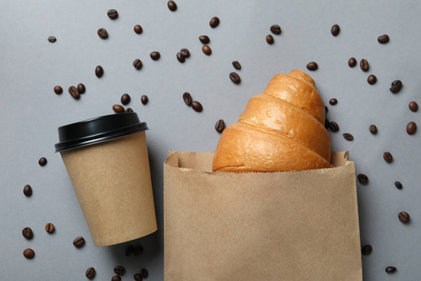 İçinde kruvasan, kağıt bardak ve gri arka planda kahve tohumu olan kağıt torba. - Fotoğraf, Görsel