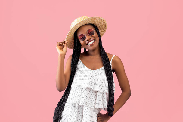 Pretty black lady with afro bunches wearing summer dress straw hat and sunglasses, smiling at camera on pink background - Foto, Bild