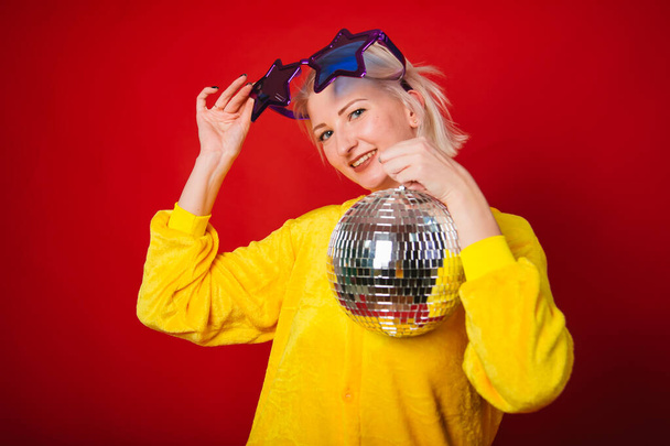 A bright girl at a party in purple big glasses in the form of stars and in yellow camisole pajamas. Funny and cheerful woman with a disco ball in her hands. Studio photo on red background - Photo, Image