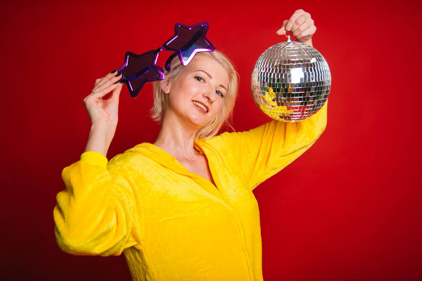 A bright girl at a party in purple big glasses in the form of stars and in yellow camisole pajamas. Funny and cheerful woman with a disco ball in her hands. Studio photo on red background - Foto, Bild