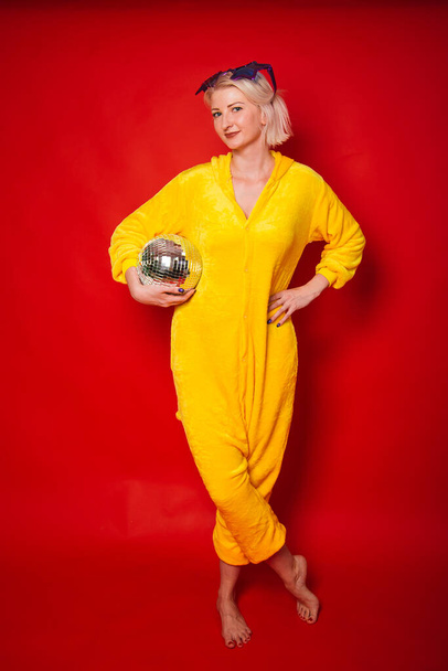 A bright girl at a party in purple big glasses in the form of stars and in yellow camisole pajamas. Funny and cheerful woman with a disco ball in her hands. Studio photo on red background - Fotó, kép