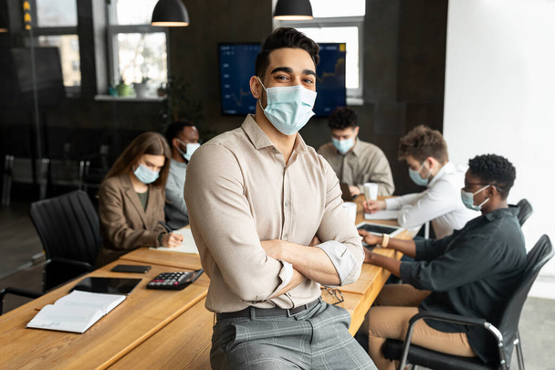 Young arab businessman in mask sitting on desk and posing - 写真・画像