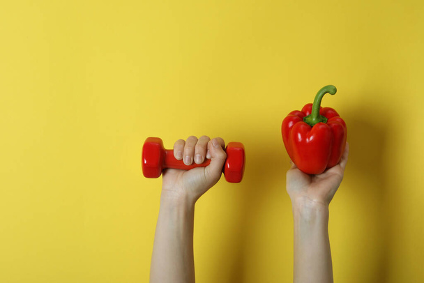 Female hands hold bell pepper and dumbbell on yellow background - Photo, Image
