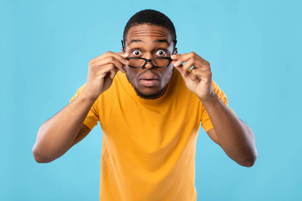 Impresionado chico africano mirando a la cámara a través de gafas, fondo azul - Foto, imagen