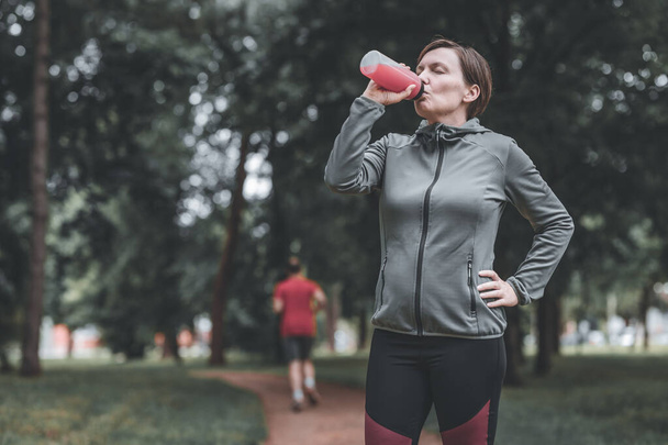 Female jogger drinking of refreshing strawberry juice in park, healthy lifestyle and recreational activity in 40s - Fotoğraf, Görsel