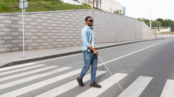 Young black visually impaired man wearing dark glasses, walking across city street, using cane - Foto, imagen