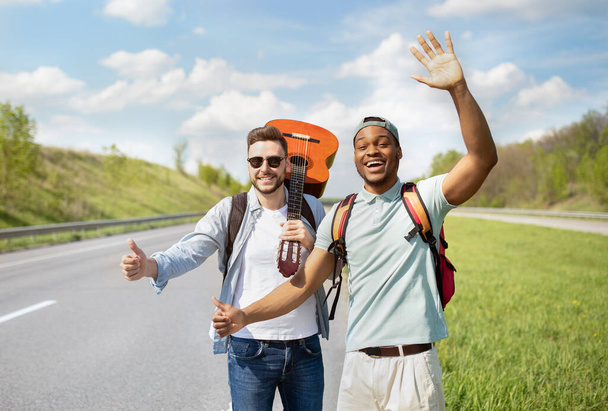 Two male friends with backpacks and guitar hitchhiking on highway, catching free ride, traveling together in summer - Foto, Imagem