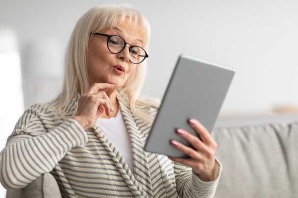 Surprised mature woman using tablet sitting on couch - Фото, зображення