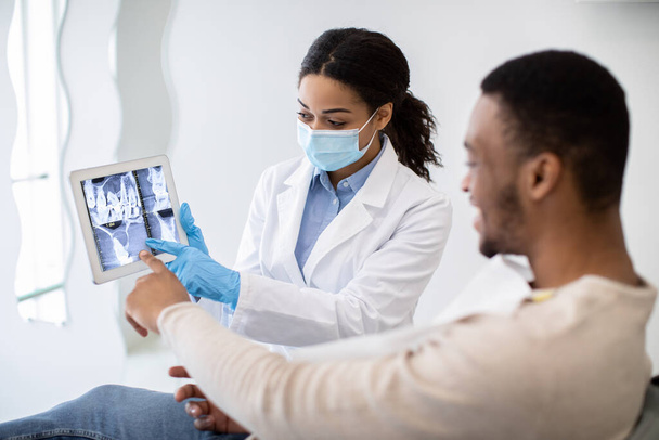 Dentiste femelle noire montrant l'image masculine de rayon X de dents de patient sur la tablette numérique - Photo, image
