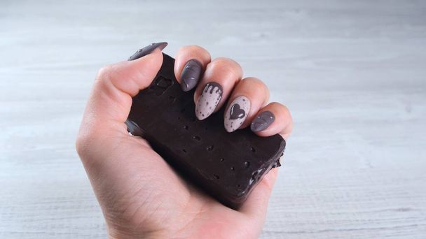Female hand with beautiful manicure nails in the form of chocolate candies, holding an unrolled bar of dark chocolate, close-up. Enjoy dessert. - Photo, Image