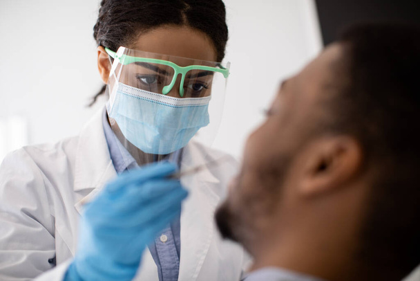 Dentista donna nera nella maschera medica e nello scudo facciale che controlla i denti dei pazienti - Foto, immagini