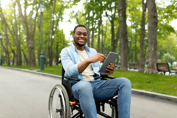 Portrét levného postiženého mladého muže na invalidním vozíku pomocí notebooku pro dálkovou komunikaci v městském parku - Fotografie, Obrázek