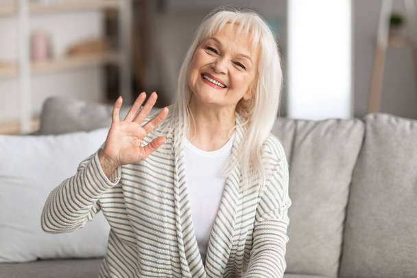 Mature woman waving at camera and smiling, sitting on sofa - Photo, Image
