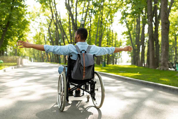 Πίσω όψη του νεαρού ανάπηρου μαύρου άνδρα σε αναπηρική καρέκλα απλώνει τα χέρια του, αισθάνεται ευτυχισμένος και ελεύθερος στο πάρκο της πόλης - Φωτογραφία, εικόνα