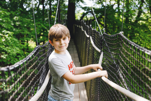 Niño preadolescente caminando por el sendero alto del dosel del árbol con pasarela de madera y cuerdas en Hoherodskopf en Alemania. Feliz niño pequeño y activo explorando el sendero de la copa de un árbol. Actividad divertida para familias al aire libre - Foto, imagen