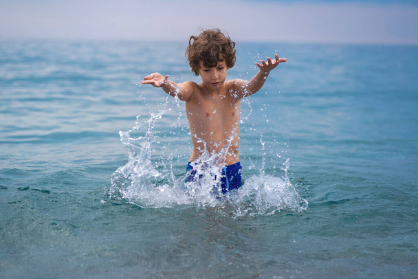 A kisfiú ugrik, és játszik a tengerbe, lövés, ahogy felbukkan a hullámok között fröccsenő víz. - Fotó, kép